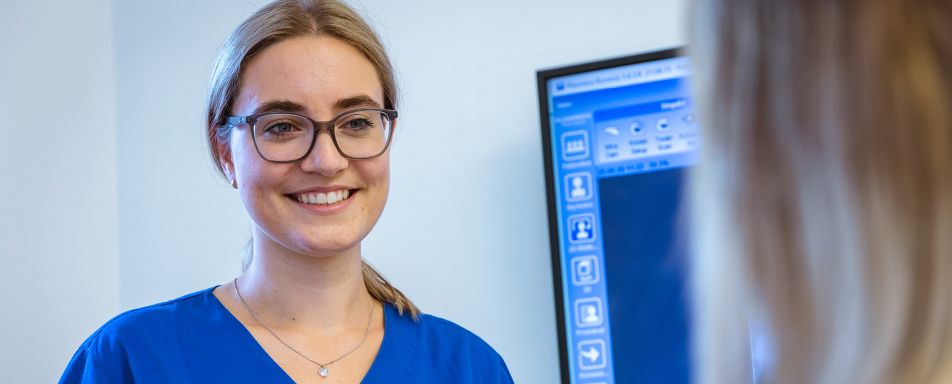 Mitarbeiterin Brille und blauer medizinischer Kleidung lächelt eine anderen Person an, die mit dem Rücken zur Kamera steht. Im Hintergrund ist ein Computerbildschirm mit verschiedenen Symbolen zu sehen.