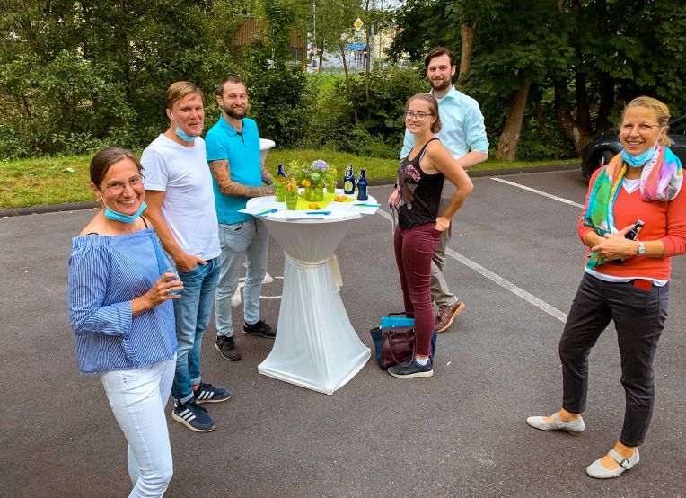 Sechs Personen stehen bei einem lockeren Treffen im Freien um einen Stehtisch mit Getränken und einem Blumenstrauß in der Mitte.