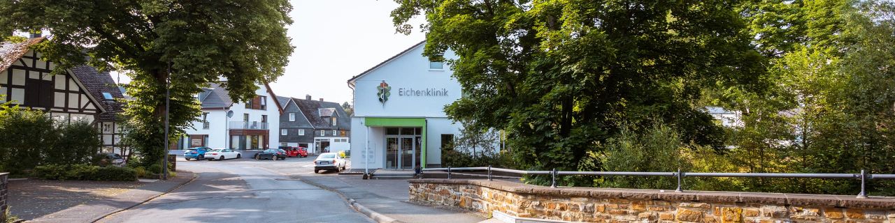 Straßenansicht der Eichenklinik. Das Gebäude ist weiß mit grünen Akzenten, umgeben von Bäumen und weiteren Häusern.