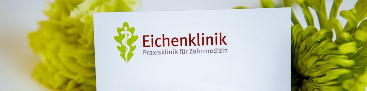 Schild der „Eichenklinik – Praxis Klinik für Zahnmedizin“ mit grünem Eichenblatt-Logo und weißem Äskulapstab. Im Hintergrund sind gelb-grüne Blumen unscharf zu erkennen.