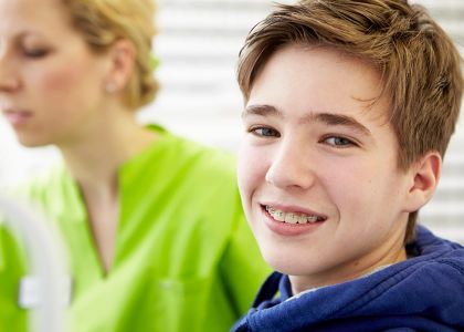 Ein braunhaariger Junge mit Zahnspange sitzt im Vordergrund, im Hintergrund eine Kieferorthopädin in grüner Kleidung.