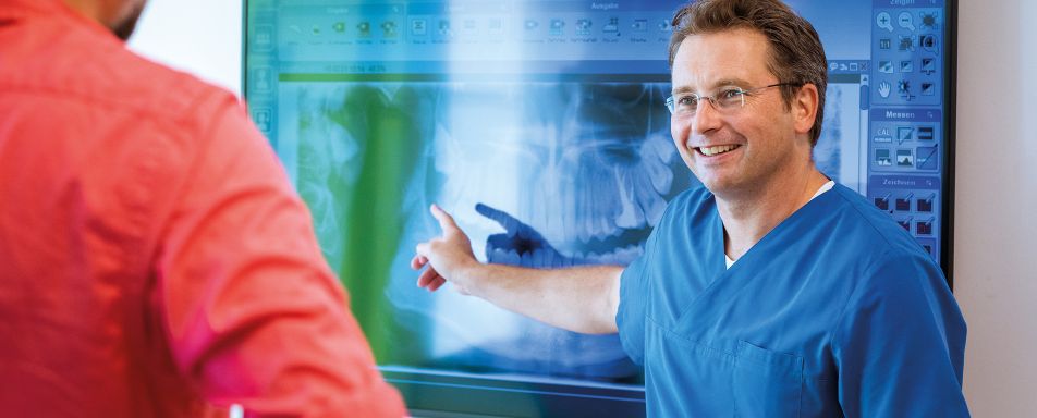 Zahnarzt in medizinischer Kleidung zeigt auf einen Bildschirm, der das Röntgenbild eines Gebisses zeigt.