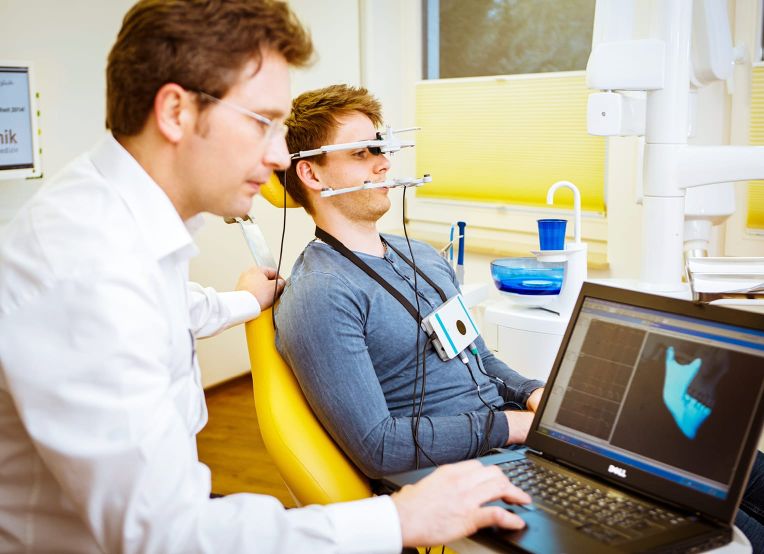 Person sitzt in einem gelben Stuhl und trägt ein Messgerät am Kopf und Gesicht zur digitalen Erfassung von Kiefer- oder Zahnstrukturen. Der Zahnarzt bedient einen Laptop, auf dessen Bildschirm ein 3D-Modell von Zähnen sichtbar.