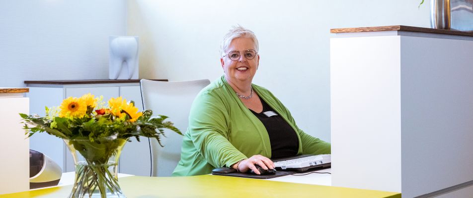 Eine freundliche Frau in grüner Kleidung sitzt lächelnd an einem Schreibtisch mit Blumen und einem Computer.