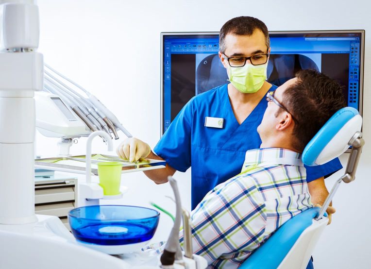 Eine Zahnarzt in blauer Kleidung, mit Maske und Handschuhen, spricht mit einem Patienten, der in einem Zahnarztstuhl sitzt. Im Hintergrund ist ein Monitor mit Zahn-Röntgenaufnahmen zu sehen, daneben zahnärztliche Instrumente.