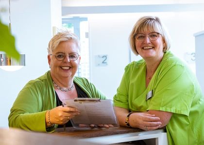 Zwei Personen – eine im Business-Outfit mit Klemmbrett, die andere in medizinischer Kleidung – die sich an einem Schreibtisch in einer hellen Arbeitsumgebung austauschen.