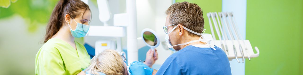 Eine Patientin wird in einem Behandlungsraum von einem Zahnarzt und einer Assistentin versorgt. Sie schaut in einen Spiegel.