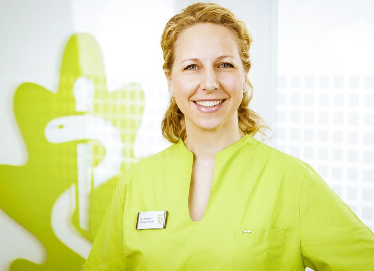 Blonde Frau mit lockigen Haaren und grünem Kasack lacht in die Kamera. Im Hintergrund ist links das Logo der Eichenklinik zu sehen.