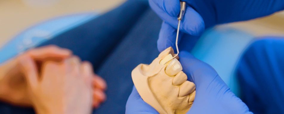 Nahaufnahme eines Zahnmodells, an dem eine Person mit blauen Handschuhen mit einem zahnärztlichen Instrument arbeitet. Im Hintergrund sind gefaltete Hände einer weiteren Person zu sehen.