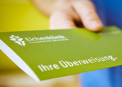 Eine Hand hält eine grüne Überweisungskarte mit der Aufschrift „Eichenklinik – Praxisklinik für Zahnmedizin“.