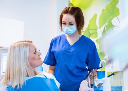 Eine Frau sitzt in einem Zahnarztstuhl, während eine weitere in blauer Kleidung, mit Maske und Handschuhen daneben steht. Im Hintergrund sind grüne Blätter als Wanddekoration zu sehen.