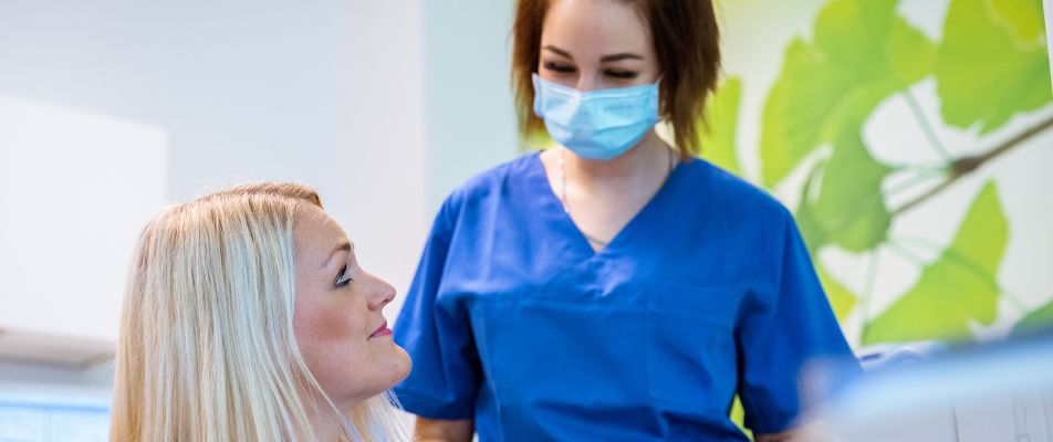 Eine Zahnärztin in blauer Schutzkleidung spricht mit einer Patientin im Behandlungsstuhl über Corona-Schutzmaßnahmen.