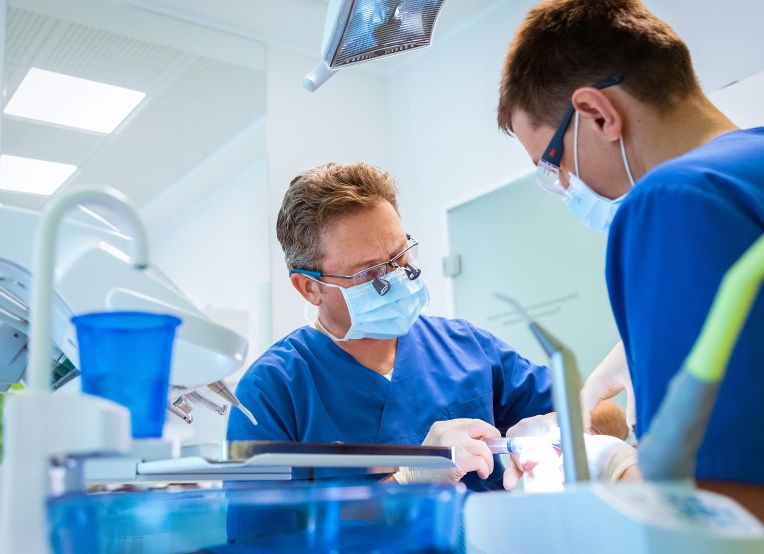 Zwei Personen in blauer Berufskleidung arbeiten gemeinsam an einer Behandlung in einer Zahnarztpraxis.