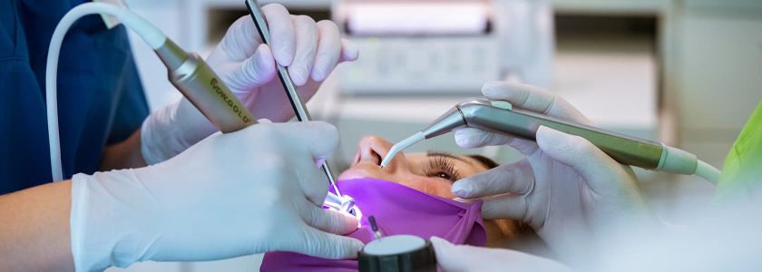 Zahnärztlichen Behandlung mit eingesetztem lila Kofferdam zur Isolierung der Zähne. Mehrere behandschuhte Hände halten zahnärztliche Instrumente wie Bohrer und Absauggerät. Im Hintergrund sind verschwommene medizinische Geräte zu erkennen.