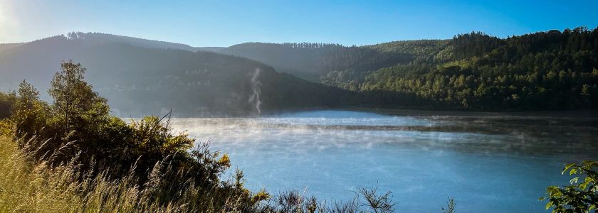 Landschaft mit einem See, umgeben von grünen Hügeln und Bäumen. Nebel liegt über dem Wasser, und Sonnenlicht fällt von links ins Bild.