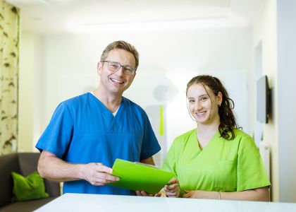 Ein Mann in blauem Kasack und eine Frau in grünem Kasack stehen in einem hellen Empfangsbereich und lächeln in die Kamera.