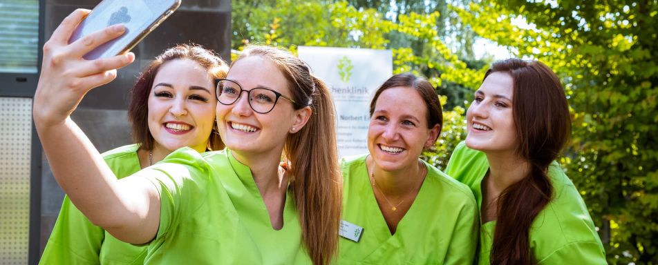 Vier Frauen in grünen Uniformen machen draußen ein Selfie. Im Hintergrund sind grüne Pflanzen und ein unscharfes Schild der Eichenklinik mit Text und Logo zu sehen.