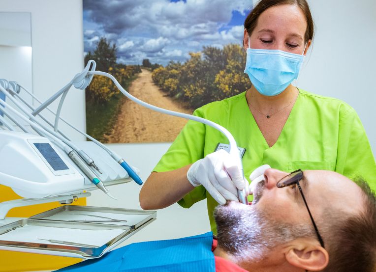 Eine zahnärztliche Fachkraft in grüner Kleidung und mit Mundschutz führt eine Behandlung an einem Mann im Zahnarztstuhl durch. Der Patient trägt eine Brille und hat eine Bart.
