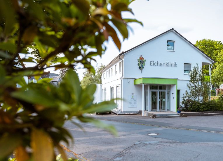 Außenansicht der Eichenklinik. Das Gebäude ist weiß mit grünen Akzenten, umgeben von Bäumen und weiteren Häusern.