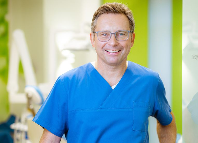 Mann mit kurzen Haaren und grauer Brille lächelt in die Kamera, er trägt einen blauen Kasack.