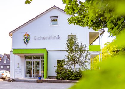 Außenansicht der Eichenklinik. Das Gebäude ist weiß mit grünen Akzenten, umgeben von Bäumen und weiteren Häusern.