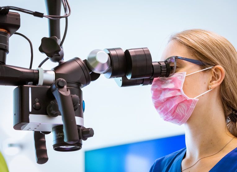 Eine Frau mit Brille und rosa Mundschutz mit weißen Mustern blickt durch ein Operationsmikroskop.