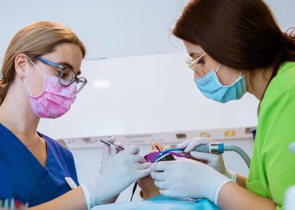 Zwei Mitarbeiterinnen führen eine Zahnbehandlung durch. Die linke trägt blaue Kleidung und eine rosa Maske, die rechte grüne Kleidung und eine blaue Maske. Beide arbeiten mit Instrumenten am Mund eines liegenden Patienten.