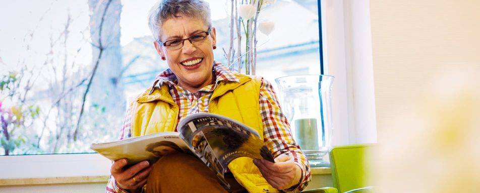 Eine Frau mit Brille und gelber Weste liest sitzend eine Zeitschrift. Im Hintergrund steht eine Vase, durch das Fenster ist ein Außenbereich zu sehen.