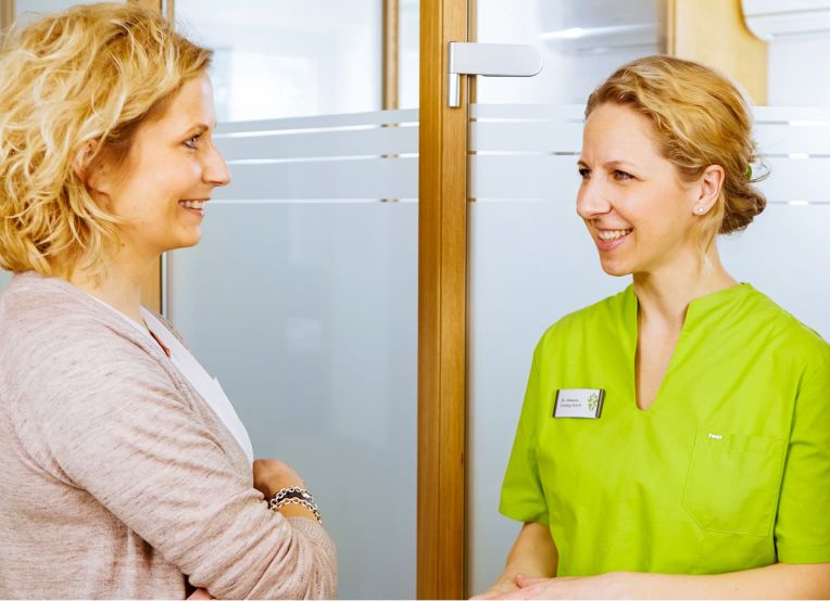 Eine Kieferorthopädin in grünem Kasack und eine blonde Frau in Alltagskleidung lächeln sich an.