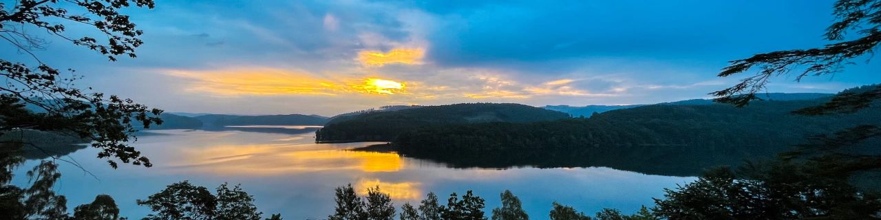 Landschaft bei Sonnenuntergang mit einem ruhigen See, umgeben von bewaldeten Hügeln. Der Himmel ist teils bewölkt und in Blau- und Orangetönen gefärbt, die sich im Wasser spiegeln.