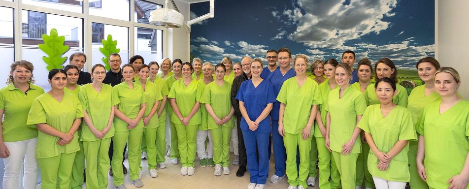 Eine Gruppe von Mitarbeitenden in grüner und blauer Berufskleidung steht gemeinsam in einem hellen Raum mit großen Fenstern - das Team der Eichenklinik.