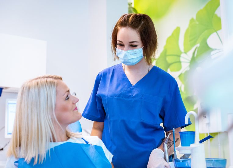 Eine ZFA in blauer Kleidung und Maske steht neben einem Patienten im Behandlungsstuhl. Im Hintergrund sind medizinische Geräte und grüne Blattdekorationen an der Wand zu sehen.