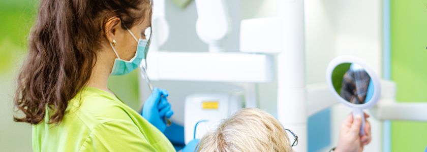 Behandlerin im grünen Kasack steht hinter einer blonden Frau, die in einen Spiegel schaut. Im Hintergrund ist verschwommen ein Behandlungsraum zu erkennen.