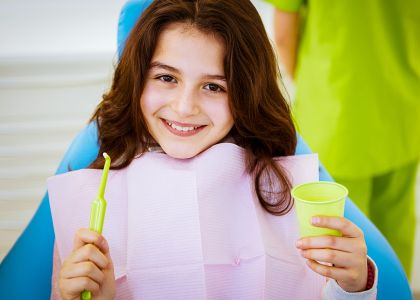 Eine braunhaariges Mädchen sitzt in einem Zahnarztstuhl mit rosa Schutzlätzchen und hält eine grüne Zahnbürste sowie einen grünen Einwegbecher. Im Hintergrund steht eine zahnärztliche Fachkraft in grüner Kleidung.