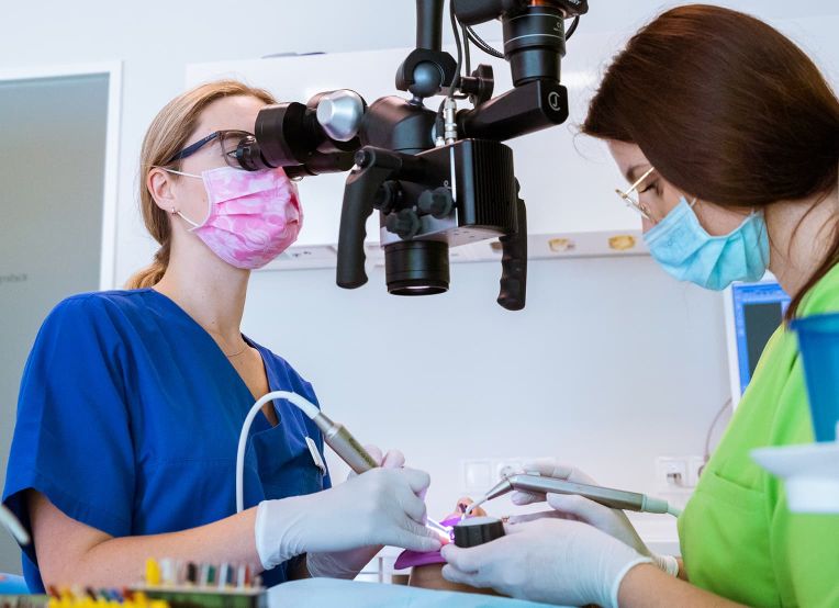 Zwei Frauen in grünen und blauen Kasacks, mit Handschuhen und Mundschutz, arbeiten an einem Operationsmikroskop.  Eine Zahnärztin blickt durch das Mikroskop, während die andere ein zahnärztliches Instrument in den Mund des Patienten hält.