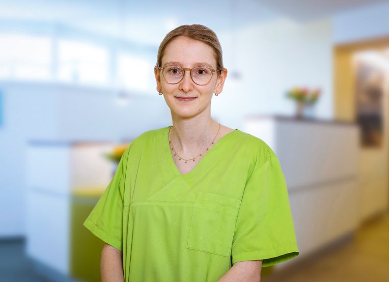 Eine Frau mit Brille in grünem Kasack steht in einem Empfangsbereich der Praxis. Im Hintergrund sind ein Tresen und Blumen zu erkennen. Die Person blickt direkt in die Kamera.