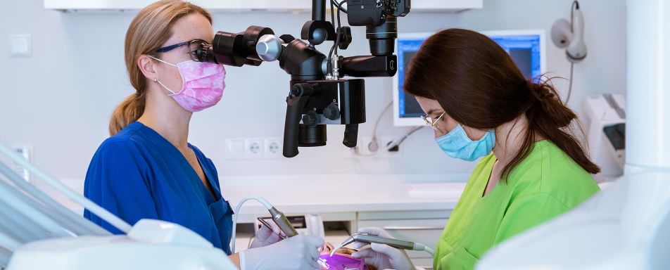 Zwei Personen in einem zahnärztlichen Behandlungsraum, eine in blauer, die andere in grüner Kleidung, beide mit Mundschutz. Eine Person arbeitet mit zahnärztlichen Instrumenten, die andere blickt durch ein Mikroskop.