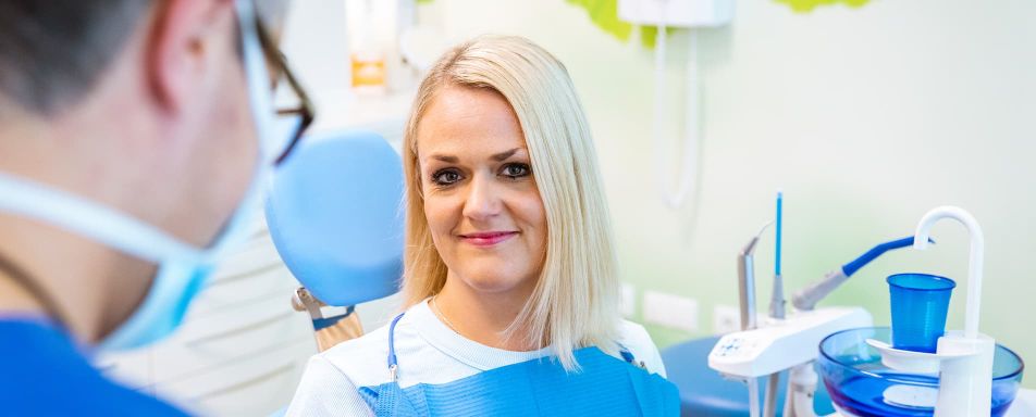 Im Fokus des Bildes sitzt eine blonde Frau auf einem Zahnarztstuhl und lächelt in die Kamera. Vorne links ist ein Mann mit OP-Maske zu sehen.