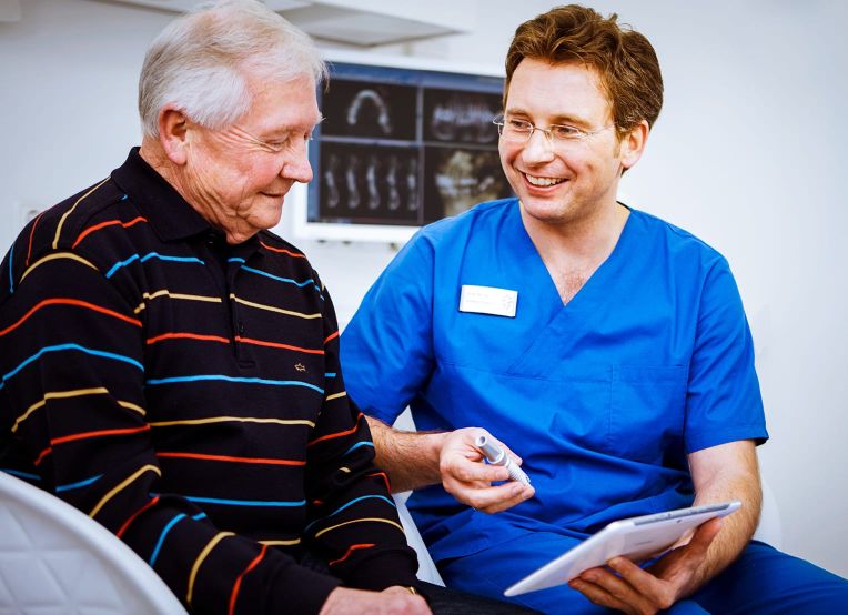 Ein Zahnarzt im blauen Karsack erklärt einem älteren Mann etwas mithilfe eines Tablets.