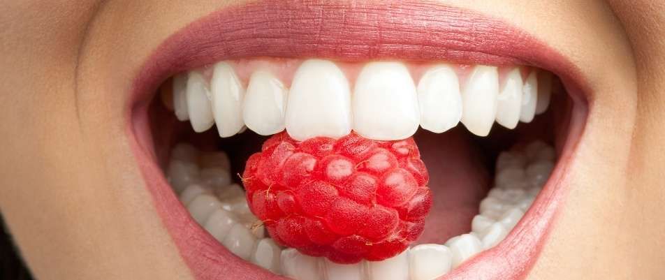 Nahaufnahme eines geöffneten Mundes mit weißen Zähnen, die eine leuchtend rote Himbeere festhalten.